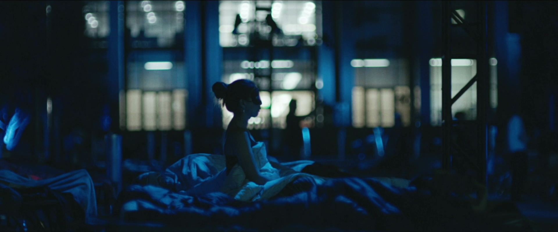 A woman sits up in bed in a dark room washed in blue night light.